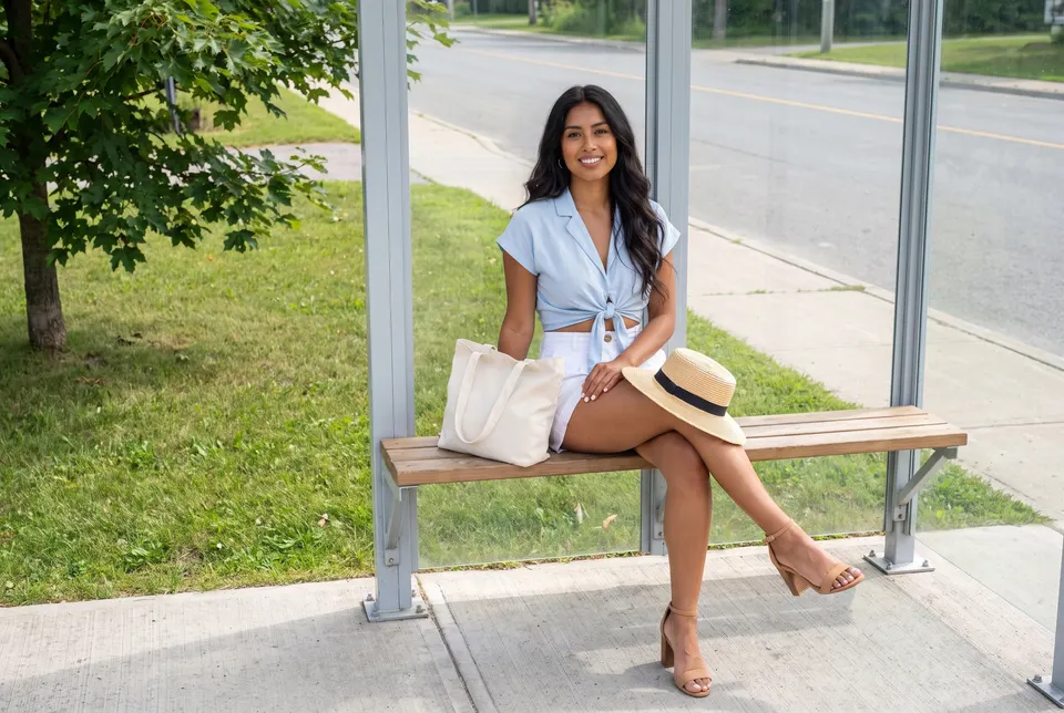The same young woman sits on a bench in a glass bus shelter on a quiet small-town street in summer, a tote bag and a straw hat beside her.